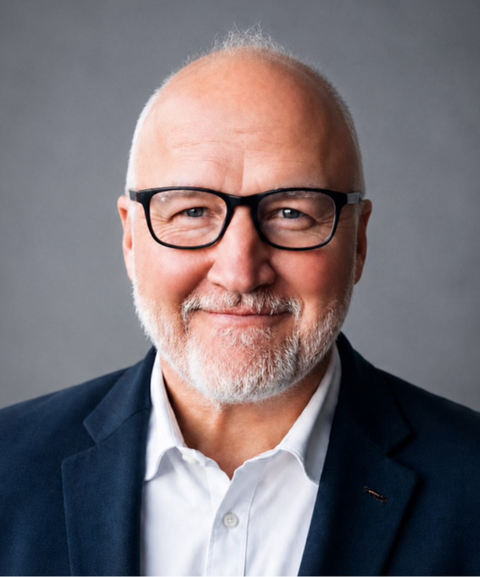 Elderly man with glasses and a white beard smiles warmly, wearing a white shirt and dark blazer, set against a gray background.