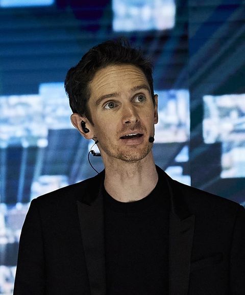 A person wearing a black suit and headset microphone speaks on stage, with a blurred digital background.