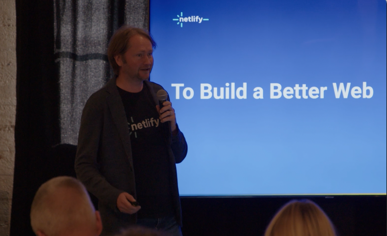 A person speaking into a microphone in front of a screen displaying "To Build a Better Web" and the Netlify logo.