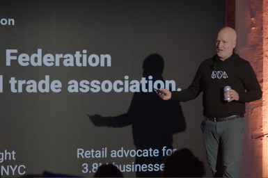 A person in a black hoodie speaks on stage, holding a drink, with a presentation screen displaying text about a trade association.