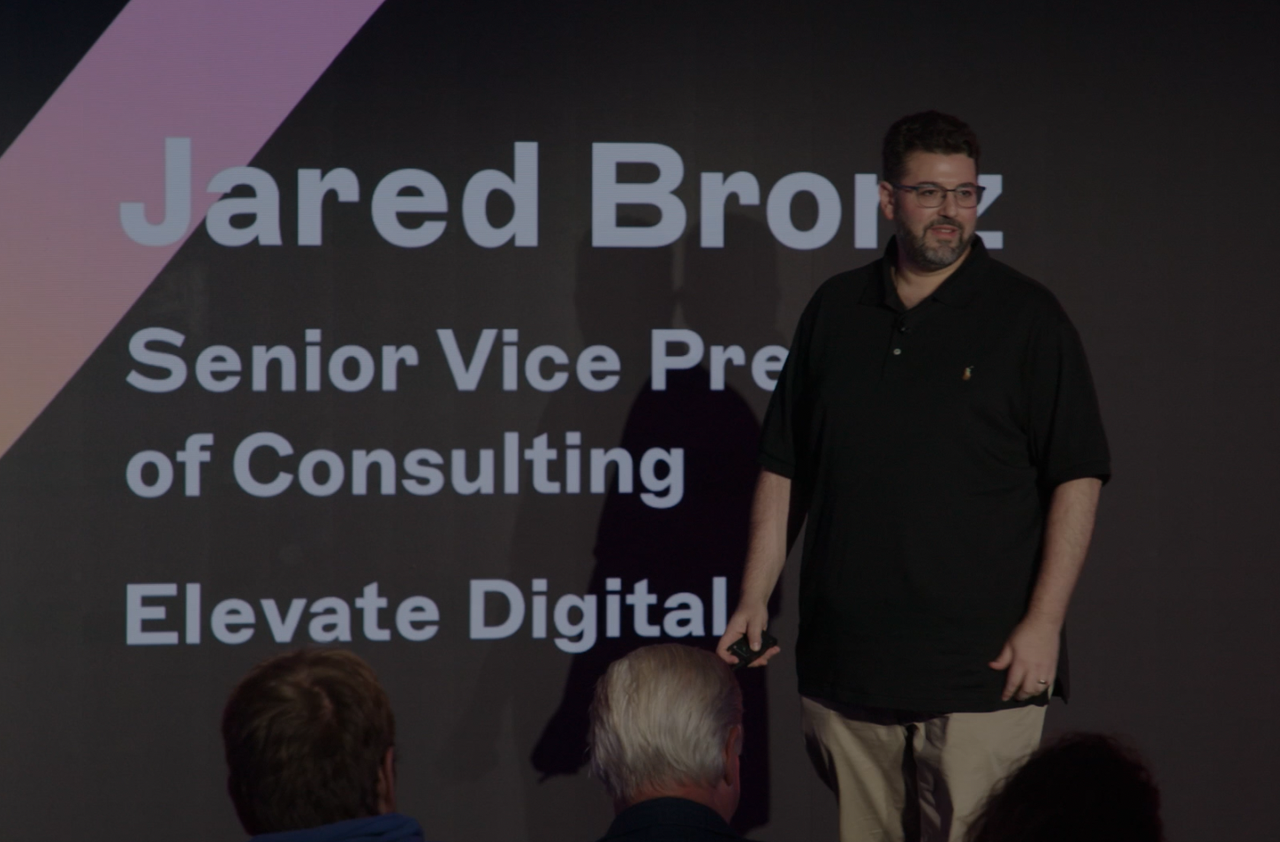A person stands on stage in front of a dark screen displaying text about a consulting role at Elevate Digital.