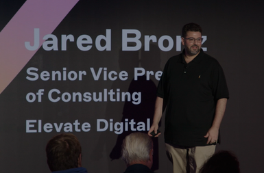 A person stands on stage in front of a dark screen displaying text about a consulting role at Elevate Digital.