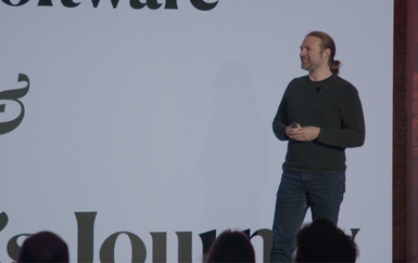 A person with long hair, wearing a dark sweater and jeans, stands on stage speaking in front of a large screen with partial text visible.