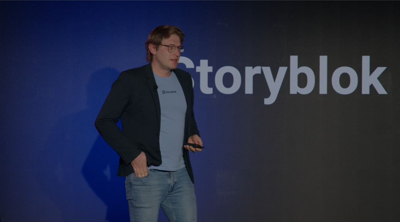 A person in a blazer and jeans speaks on stage in front of a large "Storyblok" logo.