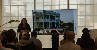 A woman presents in front of an audience, next to a screen displaying a large house and the text "About Marvin."