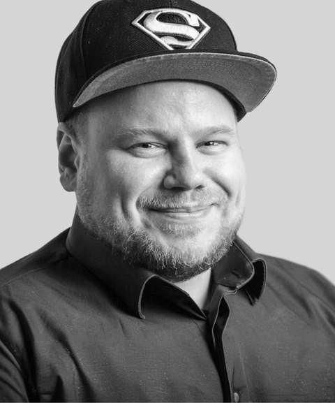 Smiling man in a black shirt and cap with a Superman logo, set against a gray background.