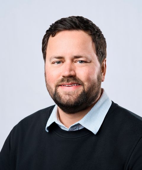 A man with short dark hair and a beard, wearing a striped shirt and a dark sweater, smiles in front of a plain light background.