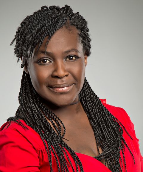 A person with braided hair smiles slightly, wearing a red top, against a neutral gray background.