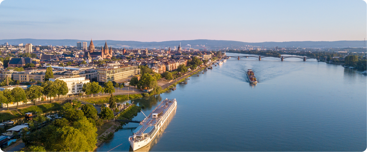 Aerial view of Mainz city with a river, trees, buildings, and a large cruise ships navigating the water, set under a clear blue sky.