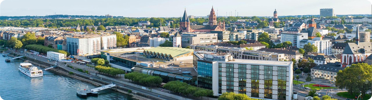 Aerial view of a city of Mainz featuring Hilton Hotel, modern and historic buildings, and lush green trees.