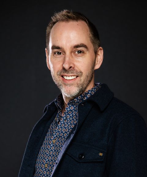 Smiling man with short hair wearing a patterned shirt and dark jacket against a dark background.