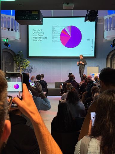 A speaker presents a pie chart on a screen to an audience, while attendees take photos with their phones.