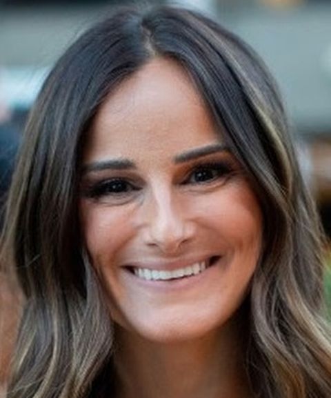 Woman with long brown hair smiling outdoors, wearing natural makeup.
