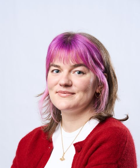 Person with pink and brown hair wearing a red sweater and gold necklace, smiling at the camera against a light background.