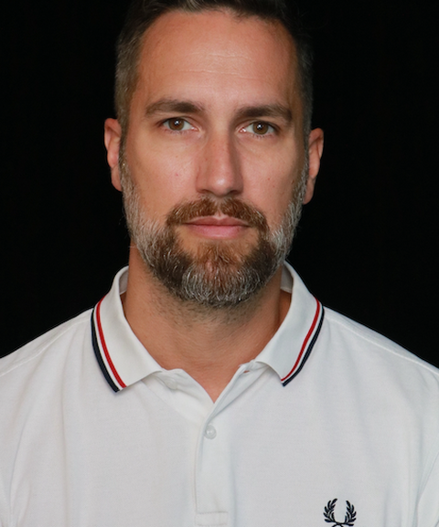 A man with a beard and short hair in a white polo shirt with black and red stripe trimming, against a black background.