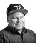 Man smiling, wearing a Superman cap and a dark shirt, in a black and white portrait.