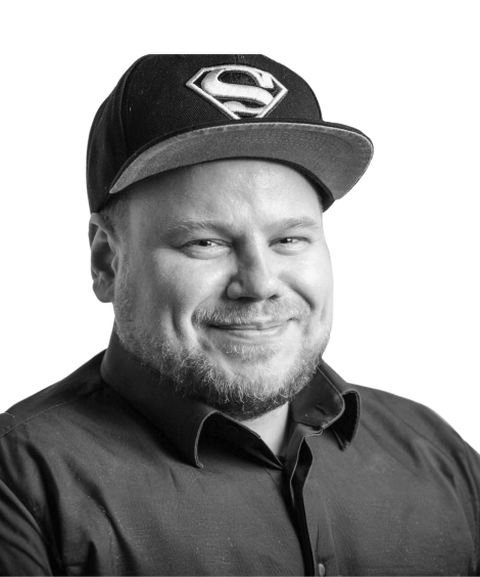 Man smiling, wearing a Superman cap and a dark shirt, in a black and white portrait.