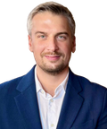A man with short hair and a beard, wearing a blue suit jacket and white shirt, smiles at the camera against a plain background.
