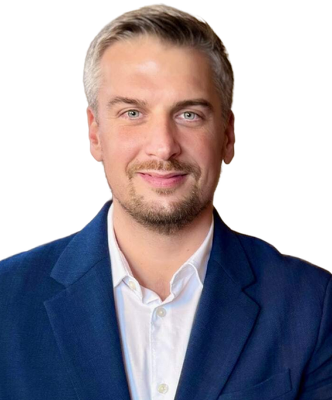 A man with short hair and a beard, wearing a blue suit jacket and white shirt, smiles at the camera against a plain background.