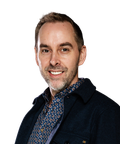A smiling man with short hair, wearing a patterned shirt and dark jacket, looking towards the camera against a plain white background.