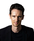 A man with short dark hair wearing a black shirt and jacket against a dark background.
