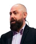 A bearded man in a suit speaking, wearing a headset microphone.