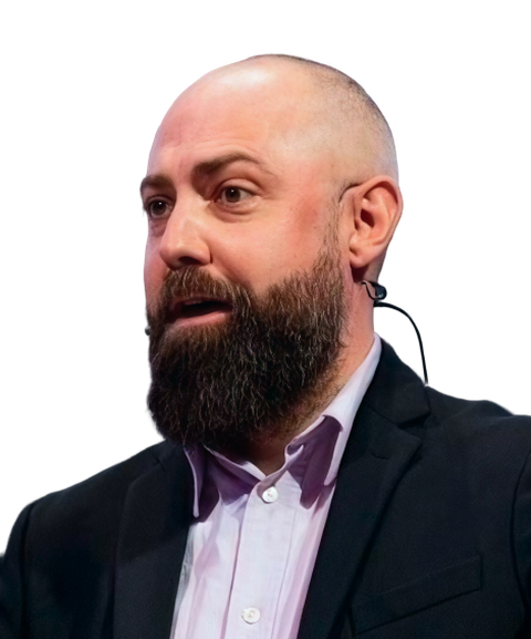 A bearded man in a suit speaking, wearing a headset microphone.
