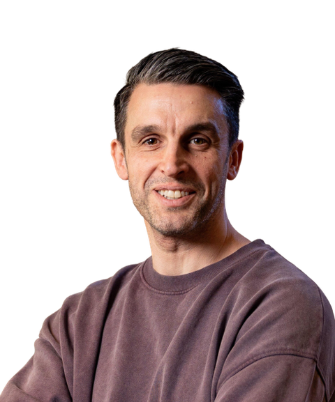 Smiling man with short dark hair and a beard, wearing a brown sweatshirt, posing against a plain white background.