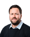 A man with short dark hair and a beard, wearing a black sweater over a light blue collared shirt, smiling against a white background.