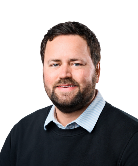 A man with short dark hair and a beard, wearing a black sweater over a light blue collared shirt, smiling against a white background.