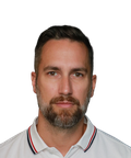 A man with short hair and a beard wearing a white polo shirt with red and black trim, looking directly at the camera against a plain background.
