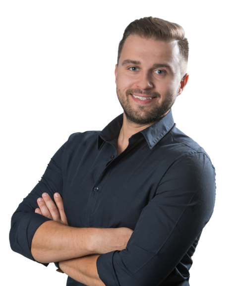 A smiling man with light brown hair and a beard stands confidently with arms crossed, wearing a dark button-up shirt against a plain background.