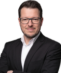 A man with glasses and a beard, wearing a black suit and white shirt, poses confidently with arms crossed.