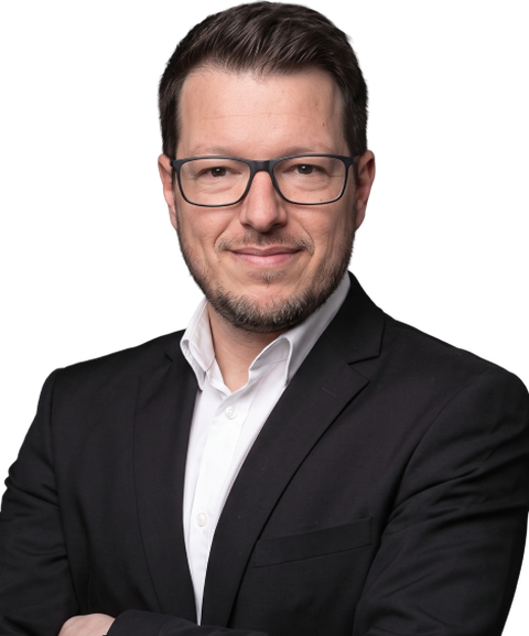 A man with glasses and a beard, wearing a black suit and white shirt, poses confidently with arms crossed.