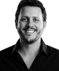 Smiling man with short curly hair and a beard, wearing a black shirt against a plain background.