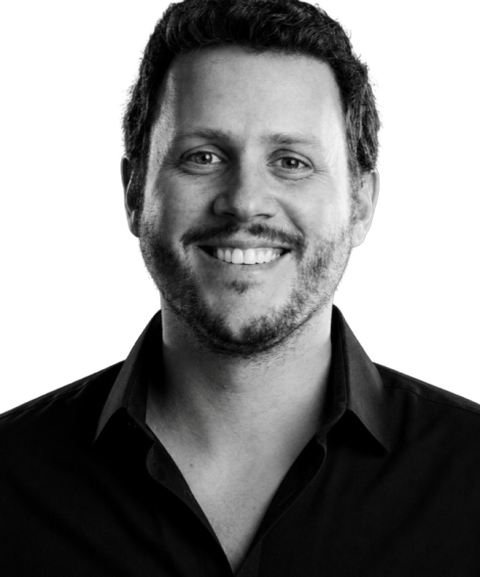 Smiling man with short curly hair and a beard, wearing a black shirt against a plain background.