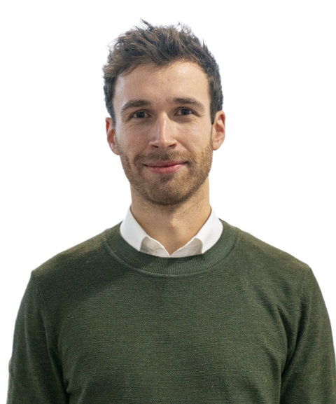 A man with short hair and a beard, wearing a green sweater over a white shirt, smiles at the camera against a plain background.