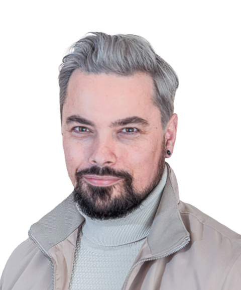 Man with gray hair and beard, wearing a light-colored turtleneck and jacket, smiling slightly against a plain background.