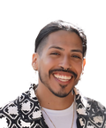 A person with long hair, a nose ring, and a mustache smiles warmly, wearing a patterned jacket and a light shirt, with a bright background.