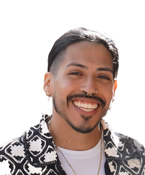 A person with long hair, a nose ring, and a mustache smiles warmly, wearing a patterned jacket and a light shirt, with a bright background.
