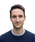 A person with short dark hair wearing a navy blue sweater, smiling slightly, set against a white background.