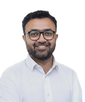 A smiling man with glasses and a beard, wearing a white shirt, stands against a plain background.