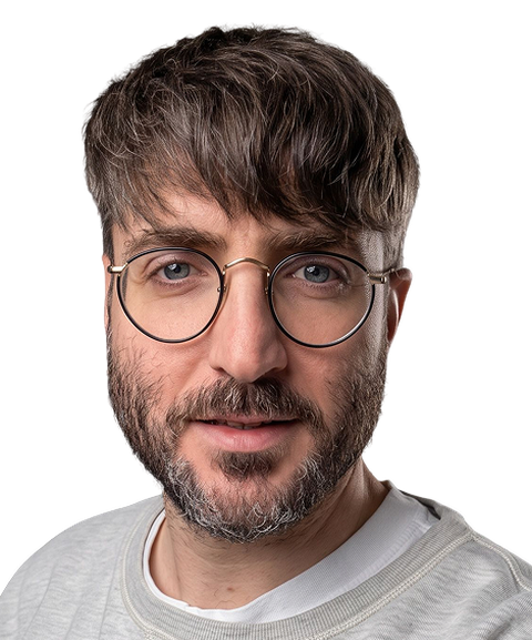 A man with short hair, wearing round glasses and a light sweater, looking directly at the camera against a plain background.