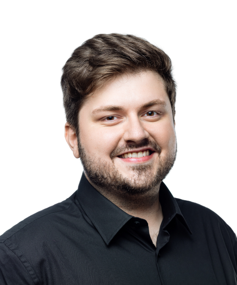 Smiling man with short brown hair and a beard, wearing a black collared shirt, against a white background.