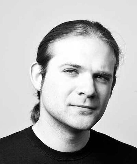 A black and white portrait of a person with long, pulled-back hair, wearing a black shirt, looking slightly to the right with a neutral expression.
