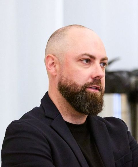 A bald man with a full beard is wearing a dark jacket. He appears to be speaking or listening attentively, with a neutral background.