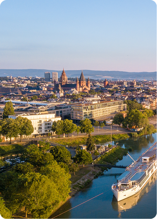 Aerial view of Mainz city with a river, trees, buildings, and a large cruise ship navigating the water, set under a clear blue sky.