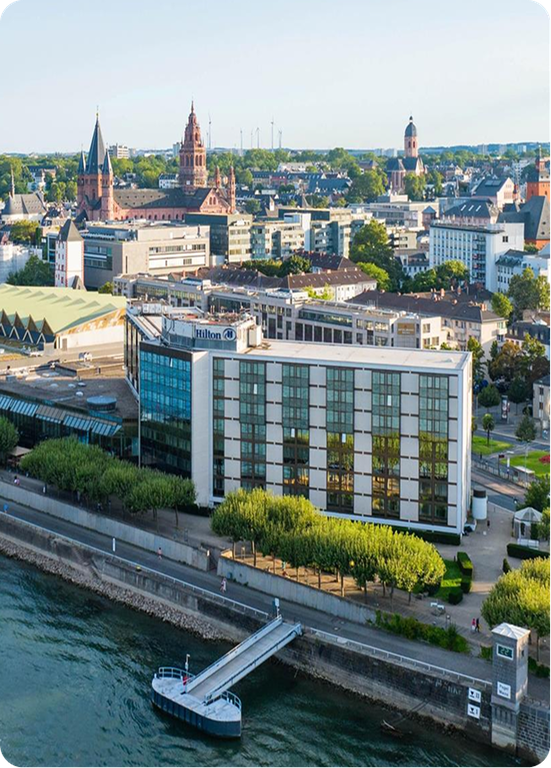 Riverside cityscape featuring a Hilton Hotel building, historic architecture with spires in the background, and a dock on the water.