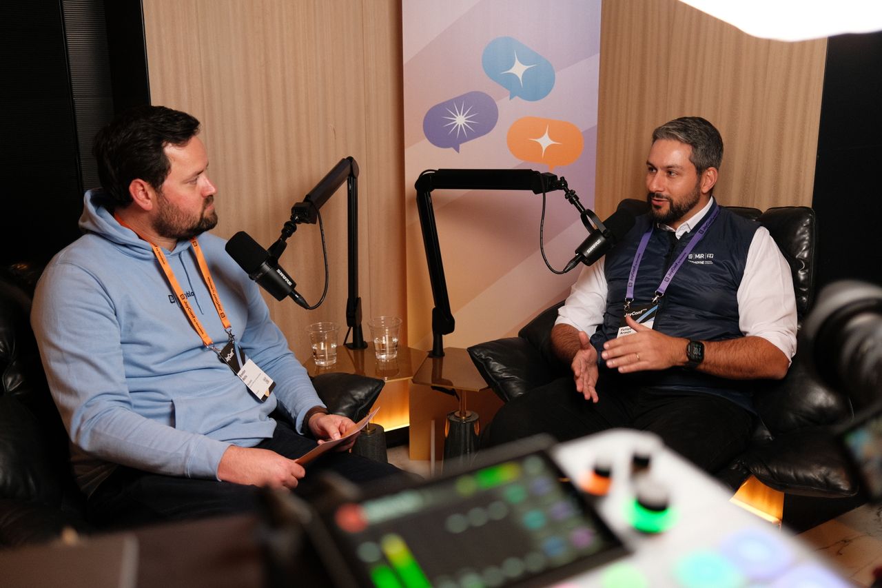 “Two men sit in leather chairs recording a podcast-style conversation. The man on the left wears a light blue hoodie and an orange event lanyard, holding a notepad while speaking into a microphone. The man on the right wears a white shirt under a navy vest with a purple lanyard, gesturing with his hands as he talks into another microphone. A sound mixer is visible in the foreground, and a banner with colorful speech bubble icons hangs behind them.