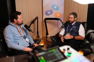 “Two men sit in leather chairs recording a podcast-style conversation. The man on the left wears a light blue hoodie and an orange event lanyard, holding a notepad while speaking into a microphone. The man on the right wears a white shirt under a navy vest with a purple lanyard, gesturing with his hands as he talks into another microphone. A sound mixer is visible in the foreground, and a banner with colorful speech bubble icons hangs behind them.